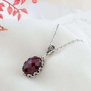 Op de afbeelding: Een zilveren ketting met een rode edelsteen hanger. De hanger is ovaalvormig en heeft een gedetailleerd zilveren frame.