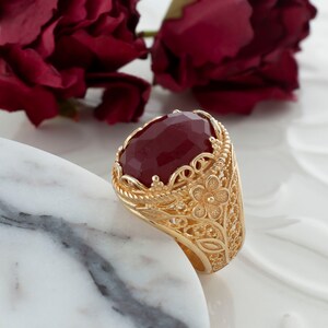 Op de afbeelding: Een gouden ring met een grote, ovale, rode edelsteen gezet in een filigraan ontwerp. De ring ligt op een wit marmeren oppervlak.