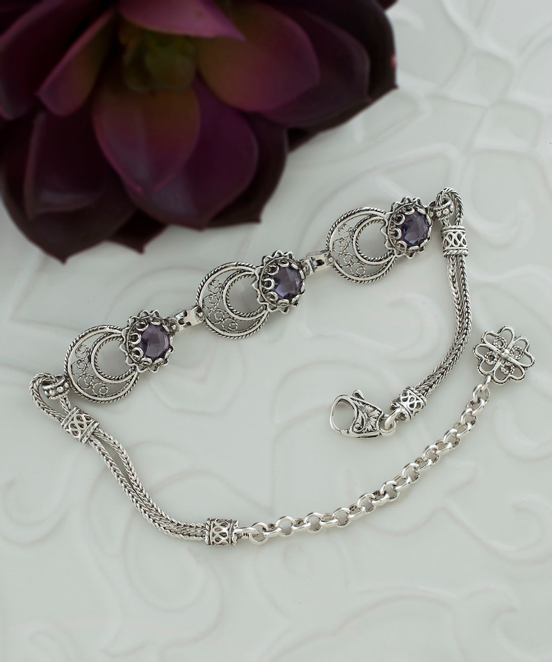 May include: A silver bracelet with three purple gemstones set in filigree circles. The bracelet has a delicate chain with a clasp and a decorative charm.