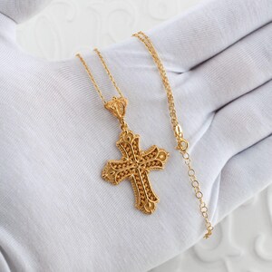 May include: A gold-colored filigree cross pendant necklace. The cross features intricate openwork detailing and is suspended from a delicate chain. The necklace is displayed on a white surface.
