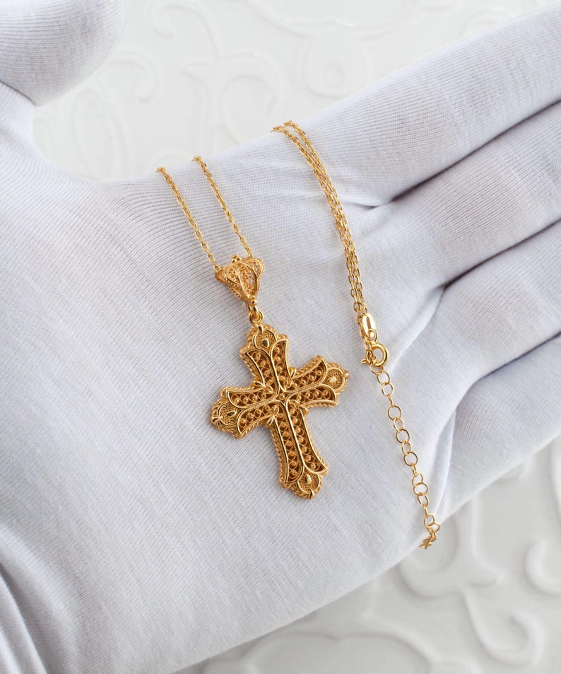 May include: A gold-colored filigree cross pendant necklace. The cross features intricate openwork detailing and is suspended from a delicate chain. The necklace is displayed on a white surface.