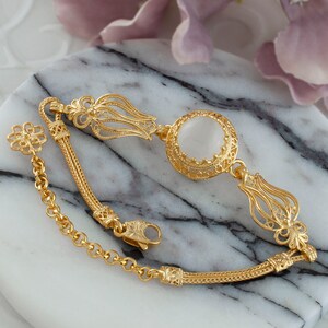 K&ouml;nnte beinhalten: Ein goldenes Armband mit einem wei&szlig;en Stein und filigranen Verzierungen. Das Armband hat ein Kettenglied und einen Verschluss.