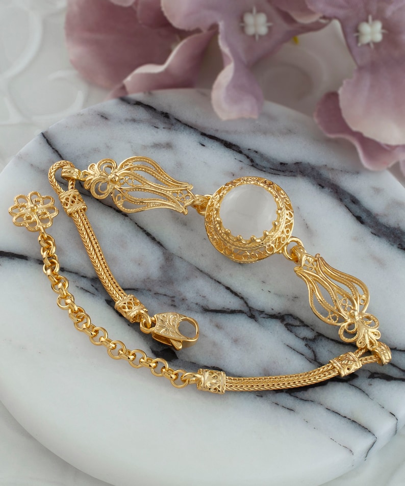 K&ouml;nnte beinhalten: Ein goldenes Armband mit einem wei&szlig;en Stein und filigranen Verzierungen. Das Armband hat ein Kettenglied und einen Verschluss.