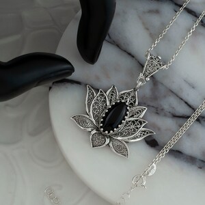 Op de afbeelding: Een zilveren filigrain lotusbloem hanger ketting met een zwarte onyx edelsteen. De ketting ligt op een wit marmeren oppervlak.
