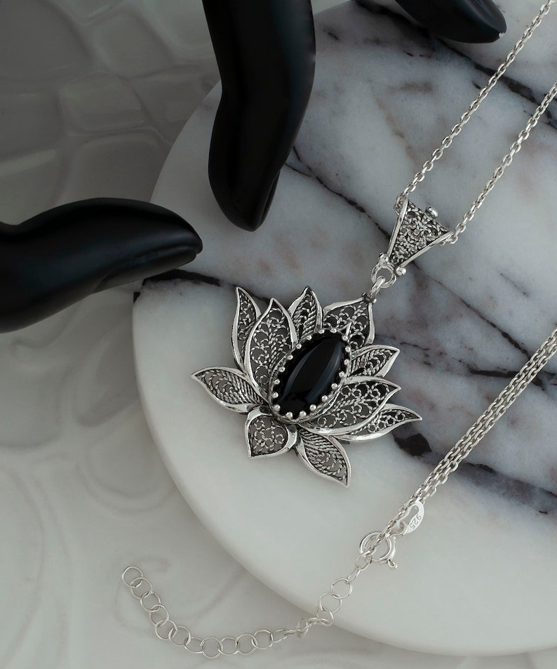 Op de afbeelding: Een zilveren filigrain lotusbloem hanger ketting met een zwarte onyx edelsteen. De ketting ligt op een wit marmeren oppervlak.