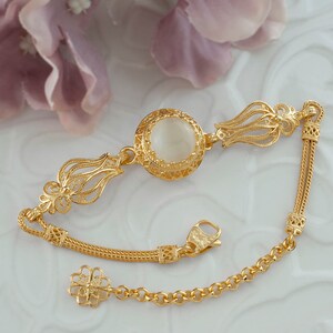 K&ouml;nnte beinhalten: Ein goldenes Armband mit einem wei&szlig;en Stein in der Mitte und filigranen Details. Das Armband hat ein Kettenglieddesign und einen Verschluss.