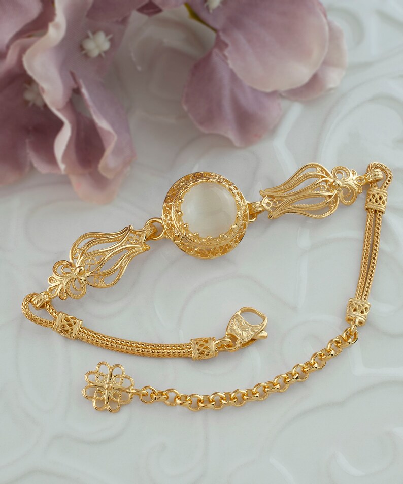 K&ouml;nnte beinhalten: Ein goldenes Armband mit einem wei&szlig;en Stein in der Mitte und filigranen Details. Das Armband hat ein Kettenglieddesign und einen Verschluss.