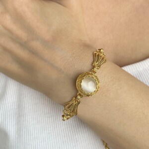 K&ouml;nnte beinhalten: Ein goldenes Armband mit einem wei&szlig;en Stein in einer filigranen Fassung. Das Armband hat eine zarte Kette und ein florales Design.