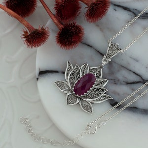 Op de afbeelding: Een zilveren ketting met een filigraan lotus hanger met een dieppaarse edelsteen. De ketting ligt op een wit marmeren oppervlak.