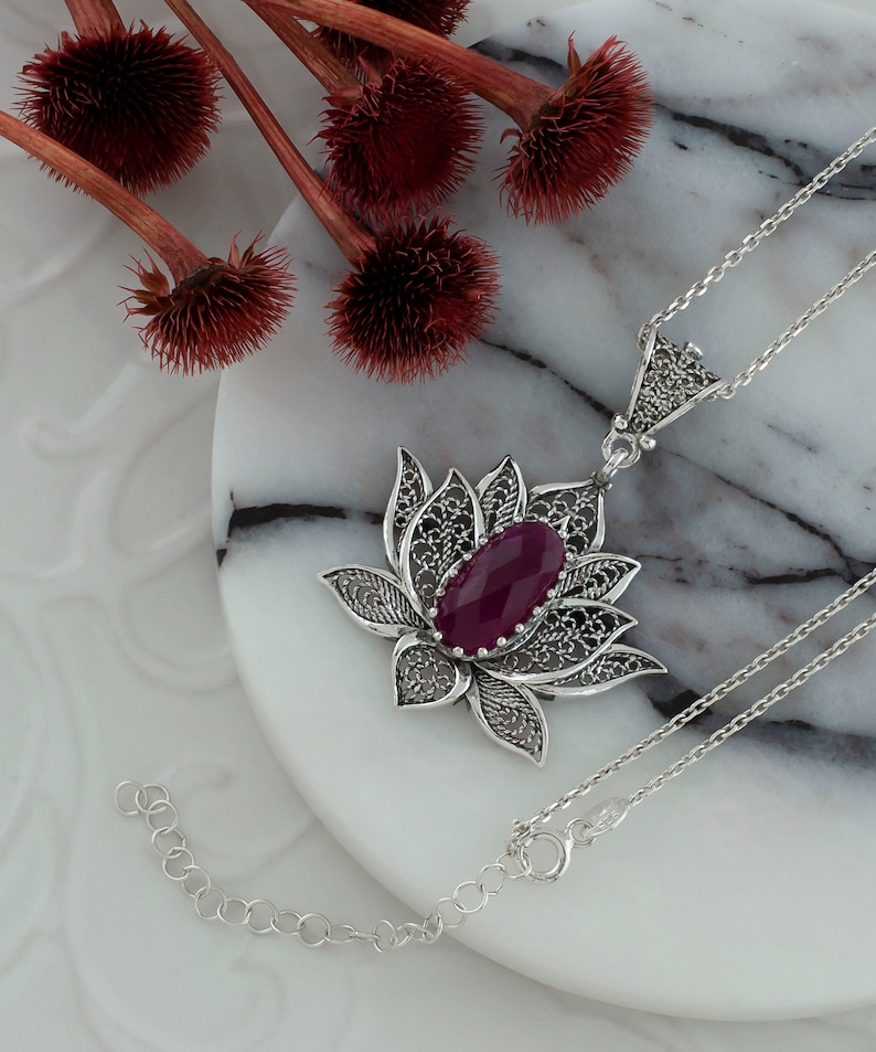 Op de afbeelding: Een zilveren ketting met een filigraan lotus hanger met een dieppaarse edelsteen. De ketting ligt op een wit marmeren oppervlak.