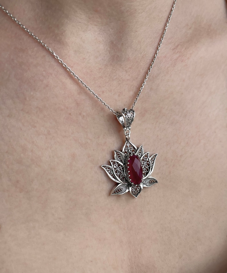 Op de afbeelding: Een zilveren ketting met een hanger in de vorm van een gedetailleerde lotusbloem met een rode edelsteen in het midden.