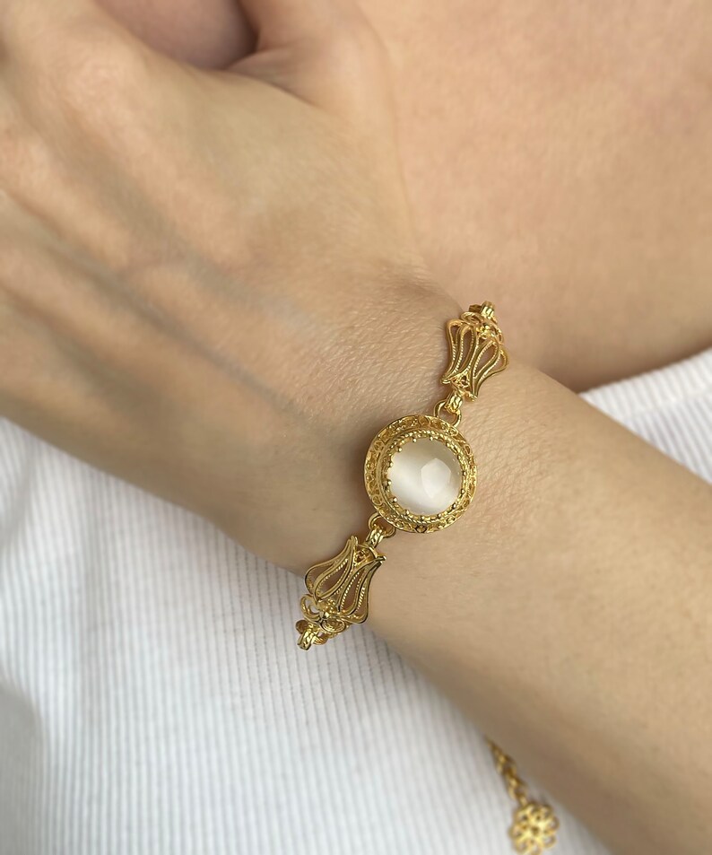 K&ouml;nnte beinhalten: Ein goldenes Armband mit einem wei&szlig;en Stein in einer filigranen Fassung. Das Armband hat eine zarte Kette und ein florales Design.