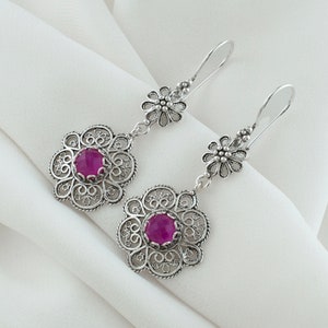 May include: A pair of silver filigree earrings with pink gemstone accents. The earrings feature a delicate floral design and dangle from a silver hook.