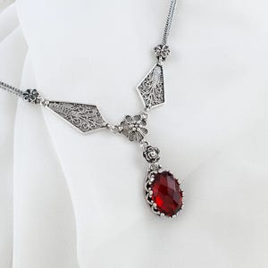 Puede incluir: Collar plateado con un colgante ovalado de piedra roja facetada. El collar tiene una cadena delicada y detalles de filigrana ornamentados en el engaste del colgante y elementos decorativos. El diseño incluye formas florales y geométricas.