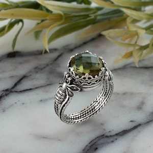 May include: A silver ring featuring a large, round, green gemstone set in a crown-like design. The band of the ring is intricately detailed with a bee design on one side.
