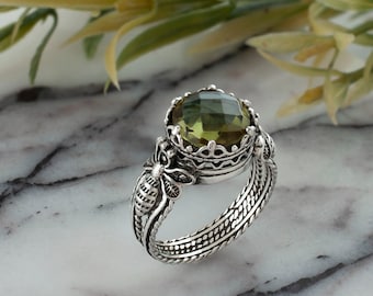 May include: A silver ring with a large, round, green gemstone set in a crown-like setting. The band of the ring is intricately detailed with a bee design on one side.