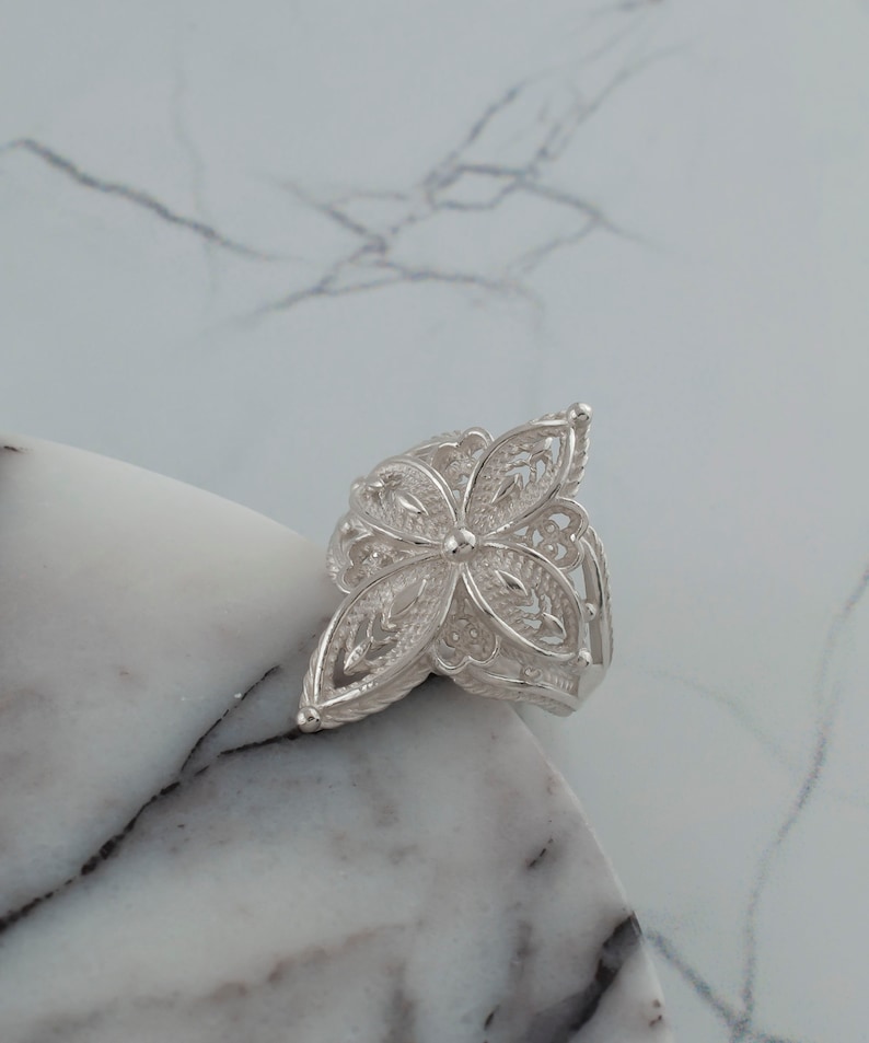 May include: A silver filigree ring with a flower design. The ring is delicate and intricate, with a floral pattern that is reminiscent of lace.