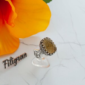May include: A silver ring with a large, oval-shaped, brown gemstone set in a filigree design. The ring is on a white display stand.