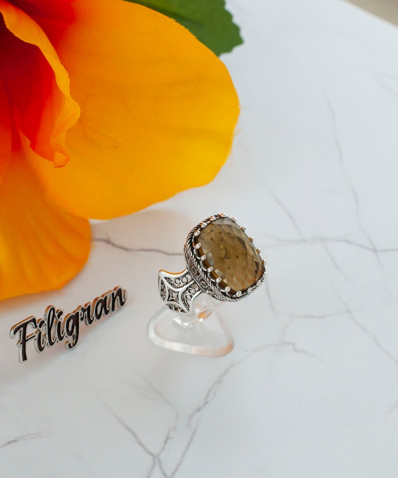 May include: A silver ring with a large, oval-shaped, brown gemstone set in a filigree design. The ring is on a white display stand.