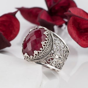 May include: A silver ring with a large, red, faceted gemstone set in a filigree design.