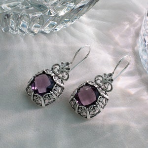 May include: A pair of silver earrings with a purple gemstone in each. The earrings have a detailed filigree design.