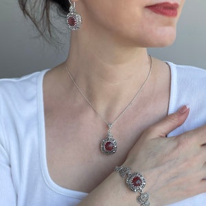 Può includere: Un set di gioielli in argento con una collana con pendente in pietra rossa quadrata e sfaccettata, orecchini abbinati e un braccialetto. I gioielli hanno un design a filigrana ornato attorno alle pietre preziose. La donna indossa un top bianco.