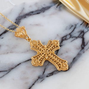 May include: A gold-colored filigree cross pendant necklace. The cross features intricate openwork detailing and is suspended from a delicate chain. The pendant is displayed on a marble surface.