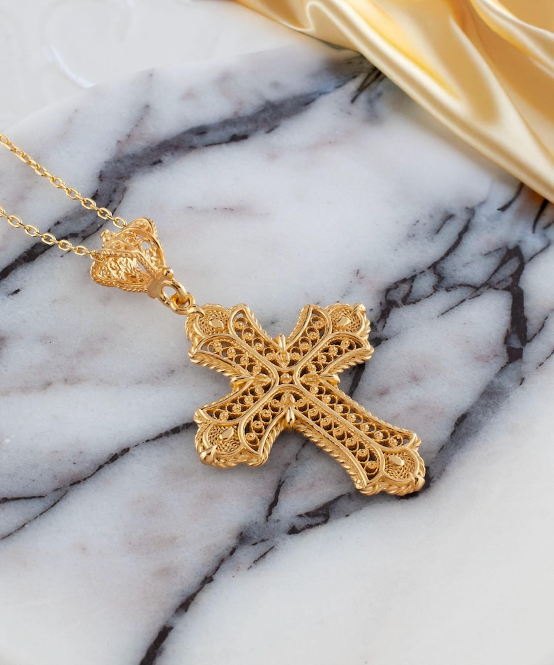 May include: A gold-colored filigree cross pendant necklace. The cross features intricate openwork detailing and is suspended from a delicate chain. The pendant is displayed on a marble surface.