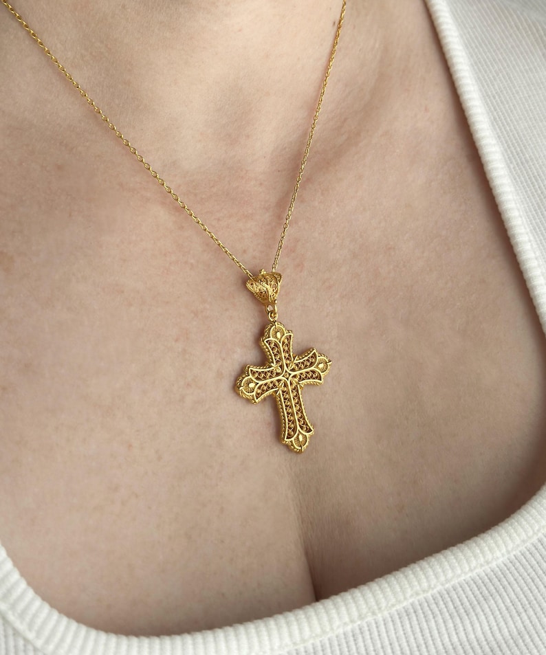 May include: A gold-colored cross pendant necklace. The cross features an ornate design with intricate detailing and is suspended from a delicate chain. The pendant is a religious symbol, often worn as a statement piece or for spiritual significance.