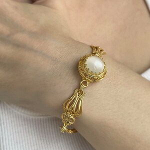 K&ouml;nnte beinhalten: Ein goldenes Armband mit einem wei&szlig;en ovalen Stein, der in einem filigranen Design eingefasst ist. Das Armband hat ein florales Motiv.