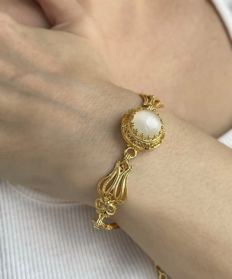 K&ouml;nnte beinhalten: Ein goldenes Armband mit einem wei&szlig;en ovalen Stein, der in einem filigranen Design eingefasst ist. Das Armband hat ein florales Motiv.
