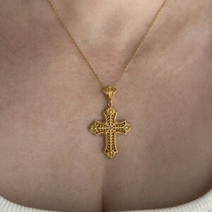 May include: A gold-colored filigree cross pendant necklace. The cross features intricate detailing and is suspended from a delicate chain. The cross is adorned with small, reddish-brown accents.