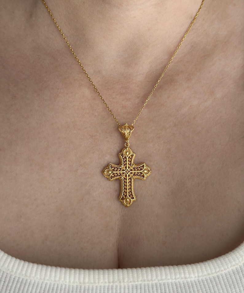 May include: A gold-colored filigree cross pendant necklace. The cross features intricate detailing and is suspended from a delicate chain. The cross is adorned with small, reddish-brown accents.