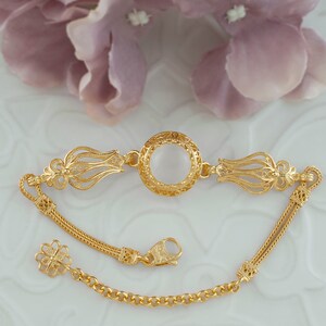 K&ouml;nnte beinhalten: Ein goldenes Armband mit einem filigranen Design und einem runden, wei&szlig;en Stein in der Mitte. Das Armband hat einen Kettenverschluss.