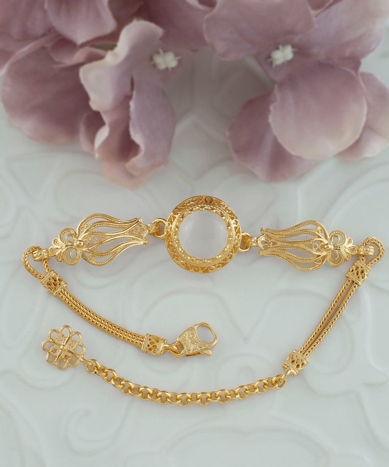 K&ouml;nnte beinhalten: Ein goldenes Armband mit einem filigranen Design und einem runden, wei&szlig;en Stein in der Mitte. Das Armband hat einen Kettenverschluss.