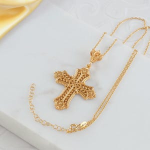 May include: A gold-colored filigree cross pendant necklace. The cross features an ornate, openwork design with intricate detailing. The necklace chain is delicate and adjustable. The pendant and chain are displayed on a white surface with a yellow fabric in the background.