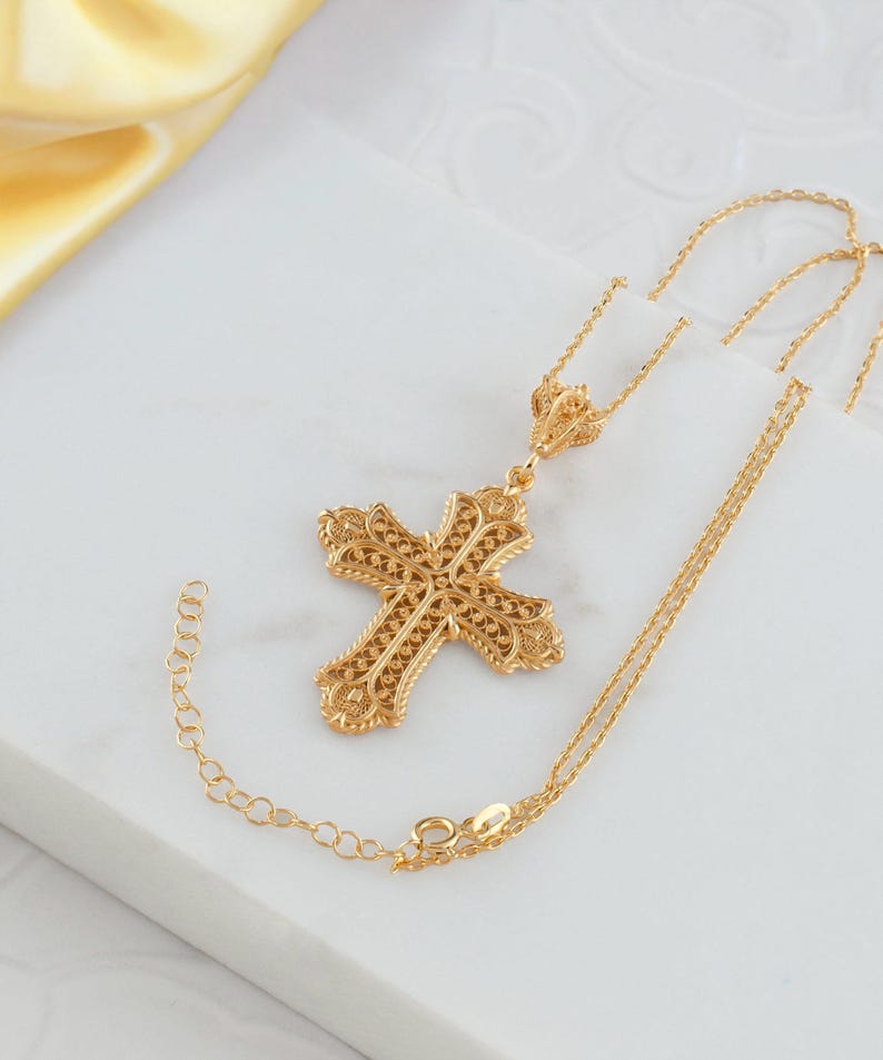 May include: A gold-colored filigree cross pendant necklace. The cross features an ornate, openwork design with intricate detailing. The necklace chain is delicate and adjustable. The pendant and chain are displayed on a white surface with a yellow fabric in the background.