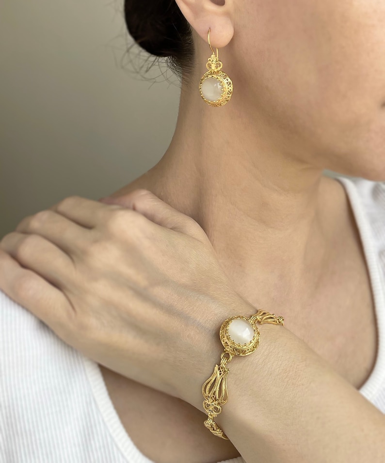 K&ouml;nnte beinhalten: Ein goldfarbenes Armband- und Ohrring-Set mit einem wei&szlig;en Stein in der Mitte jedes St&uuml;cks. Der Schmuck hat ein filigranes Design.