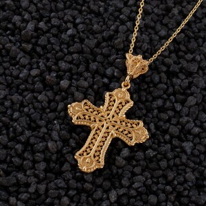 May include: A gold-colored filigree cross pendant necklace. The cross features an ornate design with intricate details and is suspended from a delicate chain. The necklace is displayed against a dark, textured background.