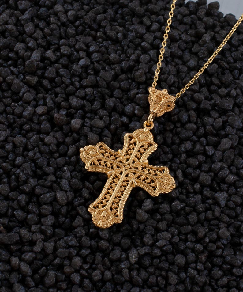May include: A gold-colored filigree cross pendant necklace. The cross features an ornate design with intricate details and is suspended from a delicate chain. The necklace is displayed against a dark, textured background.