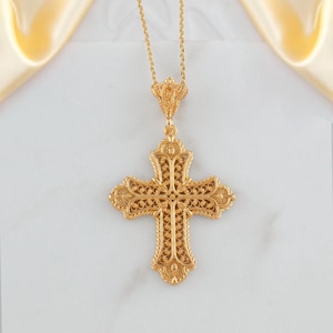 May include: A gold-colored filigree cross pendant on a delicate chain. The cross features intricate openwork and decorative detailing. The pendant hangs against a white background with a soft, golden fabric in the upper corners.