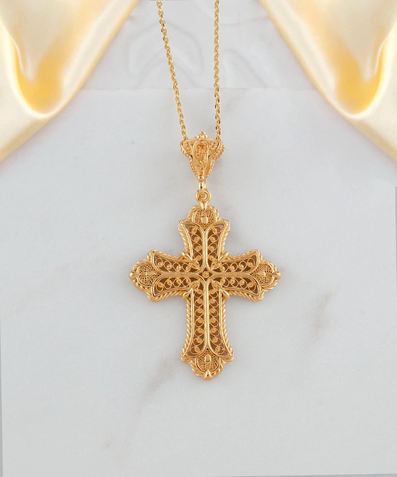 May include: A gold-colored filigree cross pendant on a delicate chain. The cross features intricate openwork and decorative detailing. The pendant hangs against a white background with a soft, golden fabric in the upper corners.