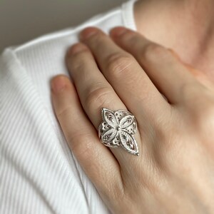 May include: A silver filigree ring with a floral design. The ring is worn on a finger.