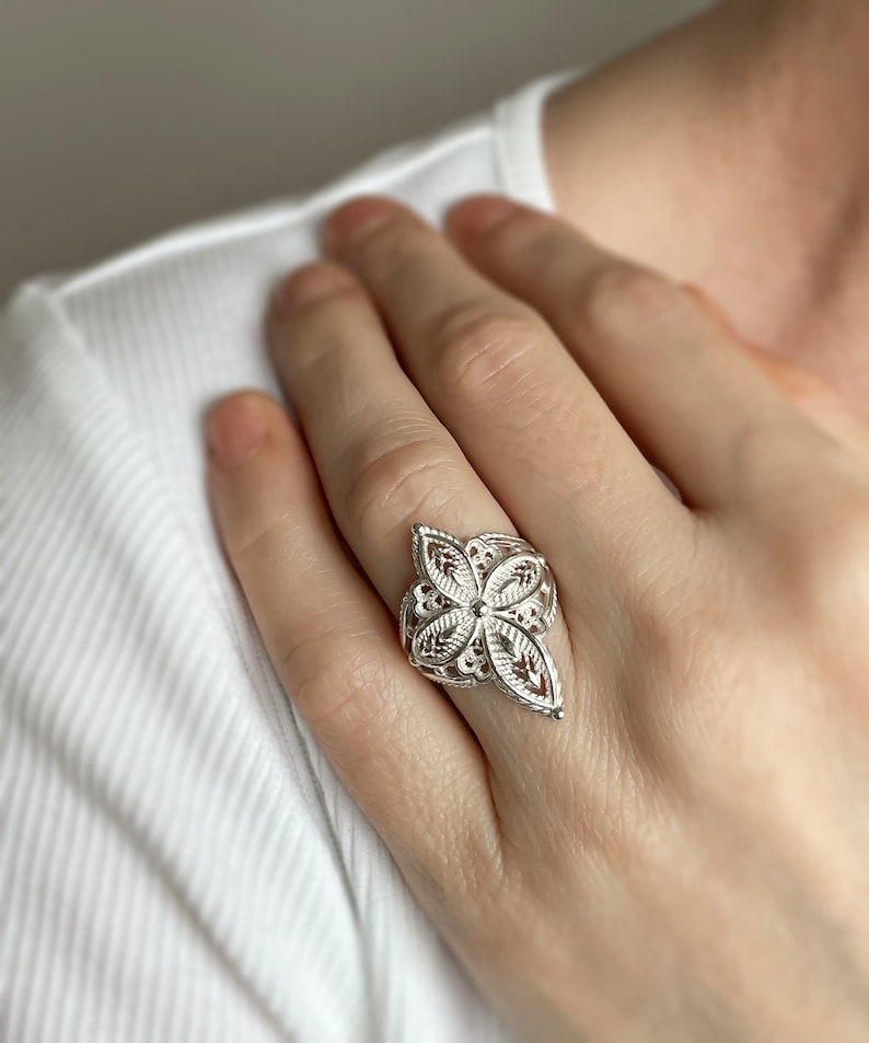 May include: A silver filigree ring with a floral design. The ring is worn on a finger.