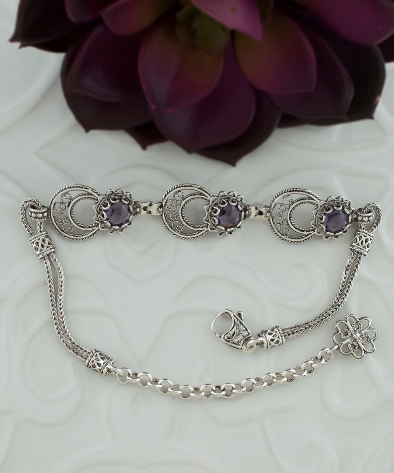 May include: A silver bracelet with three purple gemstones and a filigree design. The bracelet has a chain link closure and a decorative charm.