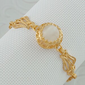 K&ouml;nnte beinhalten: Ein goldenes Armband mit einem gro&szlig;en, wei&szlig;en, ovalen Stein, der in einem filigranen Design gefasst ist.