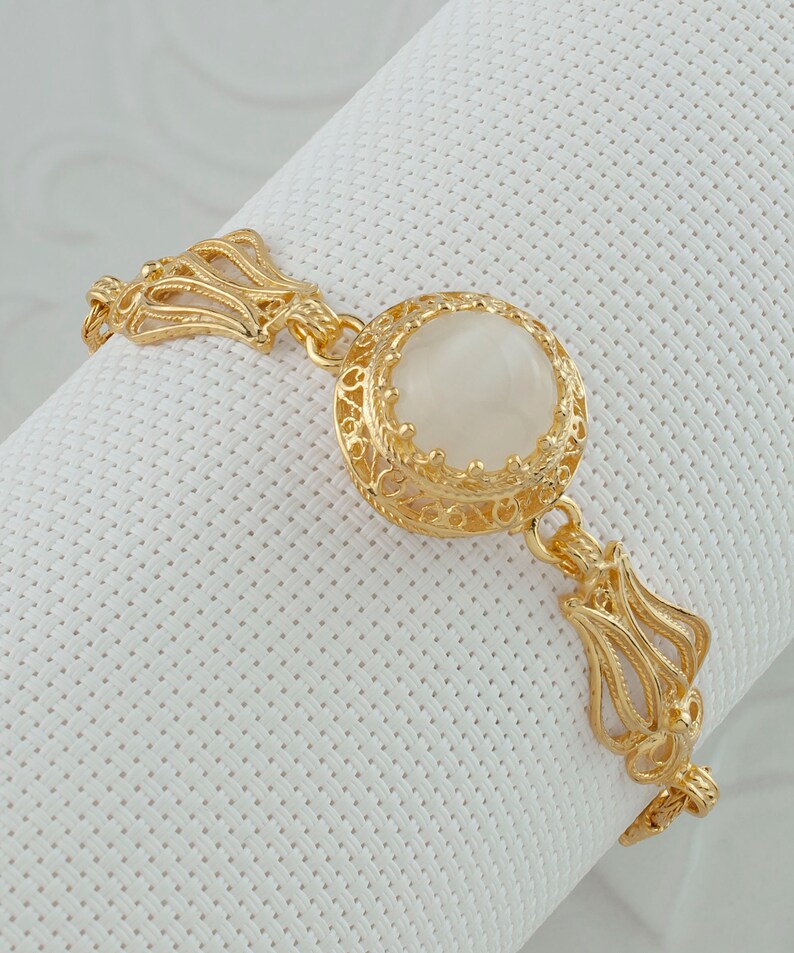 K&ouml;nnte beinhalten: Ein goldenes Armband mit einem gro&szlig;en, wei&szlig;en, ovalen Stein, der in einem filigranen Design gefasst ist.