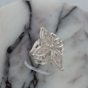 May include: A silver filigree ring with a flower design. The ring is on a white marble surface.