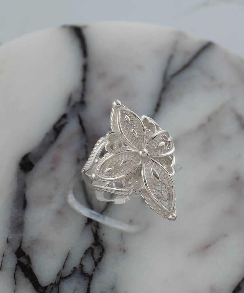 May include: A silver filigree ring with a flower design. The ring is on a white marble surface.