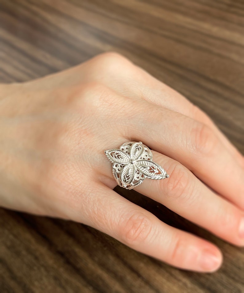 May include: A silver filigree ring with a floral design. The ring is on a person's finger.